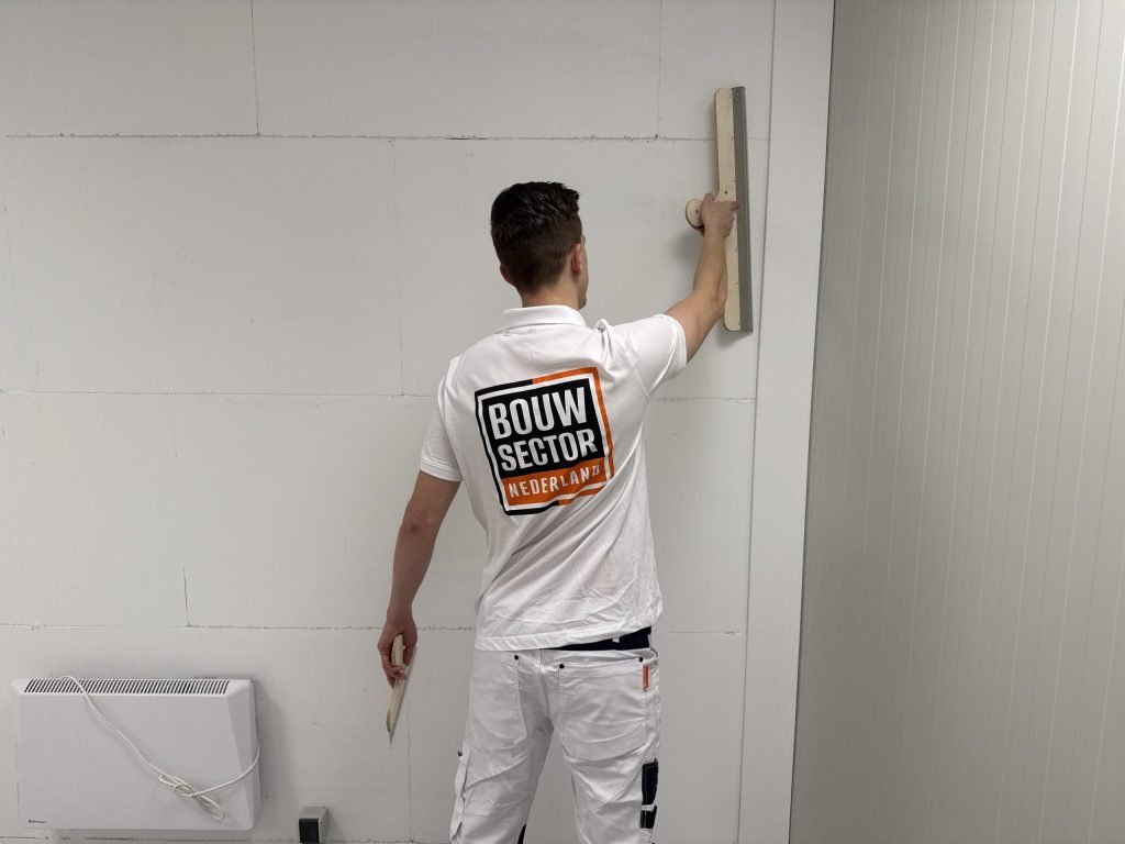 Stukadoor in wit werkpak houdt een lange spaan boven zijn hoofd tegen een muur van gipsblokken