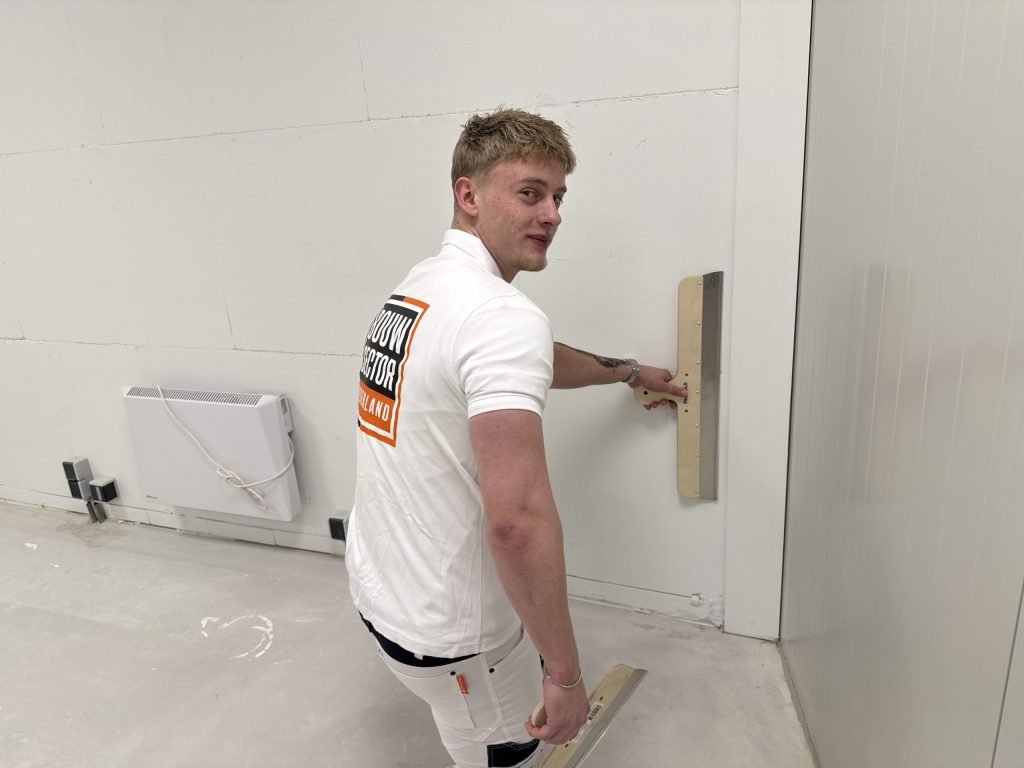 Stukadoor van Bouwsector Nederland zit op zijn knieen en strijkt het onderste deel van een muur glad met een brede spaan