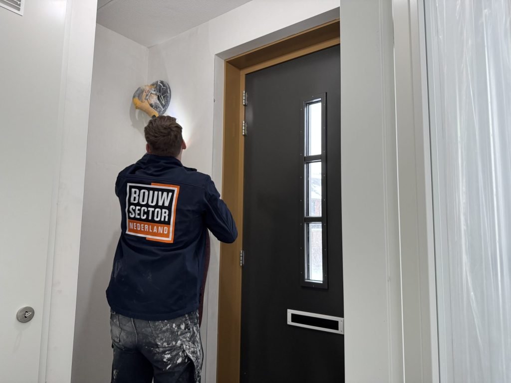 Vakman van Bouwsector Nederland in donkere werkjas schuurt met een wandschuurmachine bij de voordeur