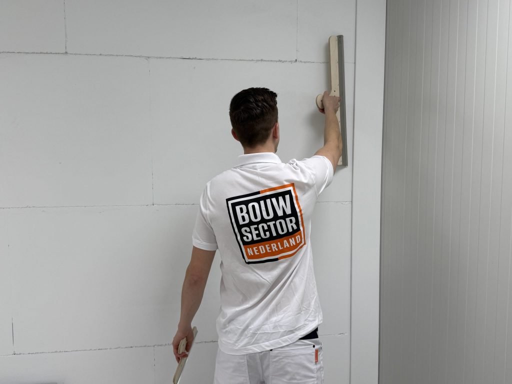 Stukadoor met het Bouwsector-logo op zijn rug zet een lange spaan verticaal tegen de wand naast een hoek
